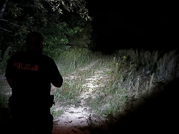 Zdjęcia przedstawiają funkcjonariusza znajdującego się w zaroślach podczas akcji poszukiwawczej za zaginioną osobą. Policjant znajduje się w umundurowaniu i oświetla miejsca zaciemnione w celu odnalezienia osoby zaginionej.