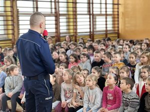 Zdjęcie przedstawia funkcjonariusza policji, który przy pomocy mikrofonu prowadzi zajęcia z dziećmi ze szkoły podstawowej. Zajęcia są na sali gimnastycznej.