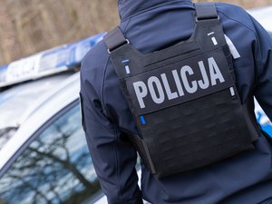 Zdjęcie przedstawia funkcjonariusza policji stojącego plecami do aparatu. Na plecach ma kamizelkę plate carrier z napisem policja.