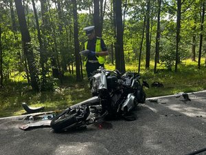 Zdjęcie przedstawia wrak motocykla w wyniku zdarzenia drogowego. Koło motocykla stoi umundurowany policjant ruchu drogowego, którzy sporządza dokumentację.