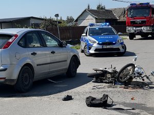 Zdjęcia przedstawiają miejsce zdarzenia drogowego, gdzie doszło do zderzenia samochodu osobowego i motoroweru. Na zdjęciach widoczny jest wóz strażacki oraz radiowóz policji.