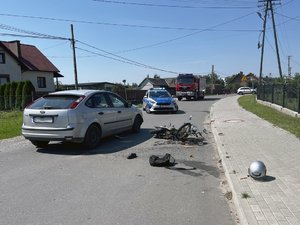 Zdjęcia przedstawiają miejsce zdarzenia drogowego, gdzie doszło do zderzenia samochodu osobowego i motoroweru. Na zdjęciach widoczny jest wóz strażacki oraz radiowóz policji.