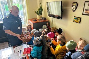 Zdjęcie przedstawia umundurowanego policjanta - I zastępcę Komendanta Powiatowego Policji w Leżajsku, który w ręce trzyma koszyk z cukierkami i częstuje nimi dzieci. Znajdują się oni w pomieszczeniu służbowym. Na zdjęciu widać grupę dzieci.