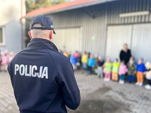 Zdjęcie przedstawia umundurowanego policjanta z napisem &quot;policja&quot; na plecach. W tle widać grupkę dzieci. Tło jest zamazane.