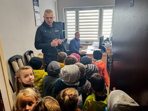 Zdjęcie przedstawia umundurowanego policjanta mówiącego do grupy dzieci. Znajdują się w pomieszczeniu służbowym. policjant skierowany jest frontem do fotografa. W tle widać innego funkcjonariusza siedzącego przed komputerem.