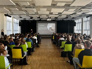 Zdjęcie przedstawia aule szkolną. W tylnej części kadr widać funkcjonariusze policji, który omawia wyświetlaną na ekranie prezentację. W przedniej części kadru po obu stronach widać siedzącą młodzież, która jest odwrócona tyłem do fotografującego.