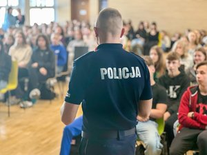 Na zdjęciu, w pierwszym planie widoczny jest umundurowany funkcjonariusz leżajskiej jednostki. Jest ubrany w tshirt policyjny. Jest odwrócony tyłem do fotografa. Na rozmazanym tle, w drugim planie widać młodzież. Kadr wykonany na auli szkolnej.