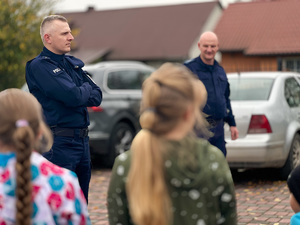 Na zdjęciu na pierwszym planie widać zamazane sylwetki dzieci, które są zamazane. Na drugim planie kadru widać dwóch umundurowanych funkcjonariuszy. W tle widać samochody.