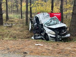 Na zdjęciu widoczny jest wrak samochodu osobowego marki Citroen C3. W tle widać drzewa, las oraz parawan straży pożarnej w kolorze czerwonym. Auto jest mocno uszkodzone.