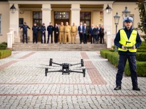 Na zdjęciu widoczny jest plac przed Muzeum Ziemi Leżajskiej. Bezpośrednio przed muzeum stoi grupa osób, w tym umundurowani funkcjonariusze Policji oraz Państwowej Straży Pożarnej. Osoby te znajdują się na ostatnim planie zdjęcia i są zamazane. Na drugim planie widoczny jest umundurowany funkcjonariusz Policji ubrany dodatkowo w żółtą, odblaskową kamizelkę z napisem policja. Funkcjonariusz Stoi frontem do aparatu i trzyma w ręku aparaturę sterującą. Na pierwszym planie, w centrum kadru znajduje się bezzałogowy statek powietrzny, który unosi się nad ziemią i jest skierowany frontem do aparatu.