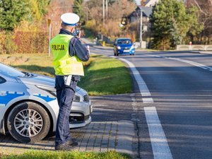 Zdjęcie przedstawia: po lewej stornie ustawiony bokiem radiowóz, a raczej jego przód. Koło niego stoi umundurowany funkcjonariusz ruchu drogowego ubrany w odblaskową kamizelkę oraz białą czapkę. Korzysta z urządzenia kontrolno-pomiarowego. W tle widać drogę, ulicę, po której porusza się w kierunku policjanta samochód osobowy.