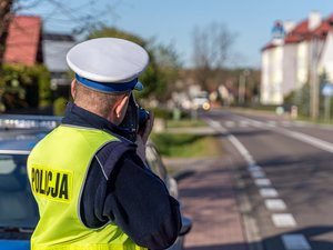 Zdjęcie przedstawia umundurowanego funkcjonariusza policji. Po jego lewej stronie widać zamazany radiowóz policji. Policjant korzysta z urządzenia kontrolno-pomiarowego. Jest ustawiony tyłem do aparatu. Drugi plan zdjęcia jest zamazany.