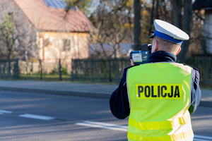 Zdjęcie przedstawia umundurowanego funkcjonariusza policji. Policjant korzysta z urządzenia kontrolno-pomiarowego. Jest ustawiony tyłem do aparatu. Drugi plan zdjęcia jest zamazany.