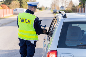 Zdjęcie przedstawia umundurowanego funkcjonariusza policji. Po jego prawej stronie widać samochód osobowy koloru srebrnego. Policjant okazuje kierującemu wynik na urządzeniu kontrolno-pomiarowym.