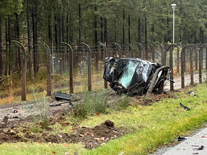 Zdjęcie przedstawia wrak samochodu marki BMW, który znajduje się na prawym boku, oparty o ogrodzenie.