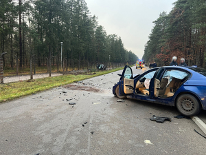 Na pierwszym planie widać uszkodzony samochód osobowy BMW w kolorze niebieskim. W tle widać odcinek drogi, oraz po lewej stornie kadru samochód, który oparty jest o ogrodzenie.