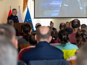 Zdjęcie przedstawia funkcjonariusza policji, stojącego w sali Muzeum ziemi Leżajskiej, który prowadzi debatę. Jest zwrócony frontem do publiczności i do frontem do aparatu. Na zdjęciu widać osoby siedzące na krzesłach. Na projektorze wyświetlany jest slajd.
