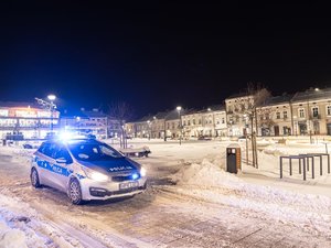 Zdjęcie przedstawia policyjny radiowóz, który skierowany jest pod kątem do obiektywu aparatu. Posiada on włączone światła oraz sygnały błyskowe. W tle widać leżajski rynek. Na zdjęciu panuje zmrok.