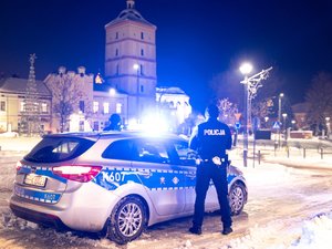 Zdjęcie przedstawia policyjny radiowóz, który skierowany jest pod kątem do obiektywu aparatu. Posiada on włączone światła oraz sygnały błyskowe. W tle widać leżajski rynek. Na zdjęciu panuje zmrok. Obok radiowozu stoi umundurowany policjant, który stoi plecami do aparatu.