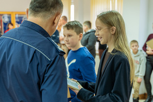 Zdjęcie przedstawia umundurowanego funkcjonariusza Policji, który wręcza nagrody laureatom konkursu &quot;Bezpieczne wakacje 2023&quot;. Na zdjęciu widać młodą dziewczynkę, która odbiera nagrodę.