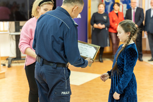 Zdjęcie przedstawia umundurowanego funkcjonariusza Policji, który wręcza nagrody laureatom konkursu &quot;Bezpieczne wakacje 2023&quot;. Na zdjęciu widać młodą dziewczynkę, która odbiera nagrodę.