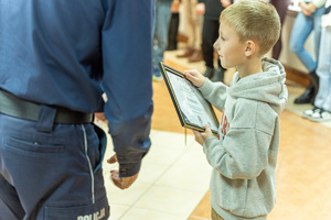 Zdjęcie przedstawia umundurowanego funkcjonariusza Policji, który wręcza nagrody laureatom konkursu &quot;Bezpieczne wakacje 2023&quot;. Na zdjęciu widać młodą dziewczynkę, która odbiera nagrodę.