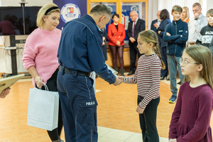 Zdjęcie przedstawia umundurowanego funkcjonariusza Policji, który wręcza nagrody laureatom konkursu &quot;Bezpieczne wakacje 2023&quot;. Na zdjęciu widać młodą dziewczynkę, która odbiera nagrodę.