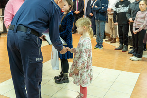 Zdjęcie przedstawia umundurowanego funkcjonariusza Policji, który wręcza nagrody laureatom konkursu &quot;Bezpieczne wakacje 2023&quot;. Na zdjęciu widać młodą dziewczynkę, która odbiera nagrodę.