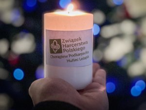 Zdjęcie przedstawia świeczkę, tak zwane Betlejemskie światełko pokoju, które znajduje się na dłoni. W tle widać zamazaną choinkę i światełka świąteczne.