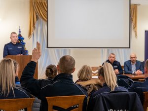 Zdjęcie przedstawia przebieg debaty społecznej pod nazwą Stop Przemocy Domowej. Na zdjęciu, na pierwszym planie widać 3 osoby, w tym 2 umundurowanych funkcjonariuszy policji i jedną osobę ubraną w odzież cywilną. Jeden z mundurowych stoi na mównicy. Pozostałe dwie osoby siedzą przy stole prezydialnym, który skierowany jest do publiczności, która na zdjęciu jest zamazana. Jedna z osób z publiczności, umundurowany funkcjonariusz Policji ma uniesioną rękę. Obok osoby stojącej na mównicy znajduje się choinka świąteczna.