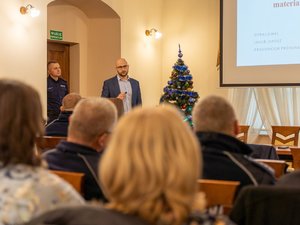 Zdjęcie przedstawia przebieg debaty społecznej pod nazwą Stop Przemocy Domowej. Na zdjęciu, na pierwszym planie widać jedną osobę ubraną w odzież cywilną. Stoi ona przy mównicy. Mównica stoi skierowana frontem do publiczności. Obok osoby stojącej na mównicy znajduje się choinka świąteczna.