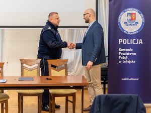 Zdjęcie przedstawia umundurowanego funkcjonariusza Policji, który wręcza drugiej osobie ubranej w odzież cywilną podziękowania. Po prawej stronie kadru znajduje się banner leżajskiej komendy.