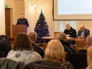 Zdjęcie przedstawia przebieg debaty społecznej pod nazwą Stop Przemocy Domowej. Na zdjęciu, na pierwszym planie widać 3 osoby, w tym 2 umundurowanych funkcjonariuszy policji i jedną osobę ubraną w odzież cywilną. Jeden z mundurowych stoi na mównicy. Pozostałe dwie osoby siedzą przy stole prezydialnym, który skierowany jest do publiczności, która na zdjęciu jest zamazana. Obok osoby stojącej na mównicy znajduje się choinka świąteczna.