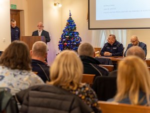 Zdjęcie przedstawia przebieg debaty społecznej pod nazwą Stop Przemocy Domowej. Na zdjęciu, na pierwszym planie widać 3 osoby, w tym jednego umundurowanego funkcjonariusza policji i dwie osoby ubrane w odzież cywilną. Jedna z osób stoi przy mównicy. Dwie osoby siedzą przy stole prezydialnym, który skierowany jest do publiczności, która na zdjęciu jest zamazana. Obok osoby stojącej na mównicy znajduje się choinka świąteczna.