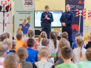 Zdjęcie przedstawia umundurowanych funkcjonariuszy Policji podczas prelekcji dla dzieci między innymi odnośnie bezpieczeństwa na przejściach kolejowych.  Funkcjonariusze stoją frontem do grupy dzieci, które siedzą na ławkach.