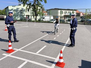 Na zdjęciu widać uczestników konkursu konkursu biorących udział w konkurencji toru przeszkód pokonywanego na rowerze oraz umundurowanych funkcjonariuszy ruchu drogowego. Zdjęcie wykonane na zewnątrz.