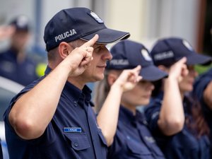 Zdjęcia przedstawiają umundurowanych funkcjonariuszy Komendy Powiatowej Policji w Leżajsku. Policjanci stoją na zewnątrz i oddają honory, przykładając  2 palce prawej dłoni do czapka z daszkiem, na którym znajduje się wyhaftowany Polski Orzełek. W tle widoczne są radiowozu oznakowane i budynek komendy.