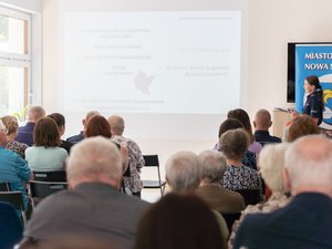 Zdjęcie przedstawia umundurowanego funkcjonariusza, znajdującego się w pomieszczeniu zamkniętym. Skierowany jest frontem do aparatu. Przed nim znajdują się osoby, które na zdjęciu są zamazane.