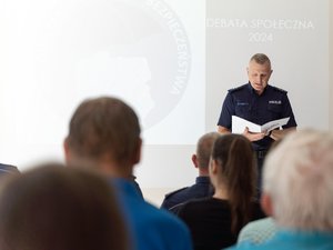 Zdjęcie przedstawia umundurowanego funkcjonariusza, znajdującego się w pomieszczeniu zamkniętym. Skierowany jest frontem do aparatu. Przed nim znajdują się osoby, które na zdjęciu są zamazane.