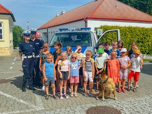 Zdjęcie przedstawia grupę dzieci na tle zaparkowanego, oznakowanego radiowozu. po lewej stronie widać trójkę policjantów, umundurowanych. W środku kadru znajduje się pies służbowy Dżina owczarek belgijski. Zdjęcie zostało wykonane na zewnątrz.