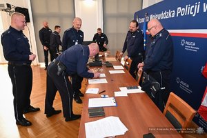 Zdjęcia przedstawiają umundurowanych funkcjonariuszy Policji w trakcie przebiegu eliminacji do ogólnopolskiego konkursu służby dyżurnej. Na zdjęciach widać zmagania policjantów podczas testów i zadań praktycznych. Wszystko odbywa się w zamkniętej sali konferencyjnej.