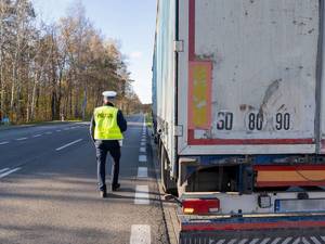 Zdjęcie przedstawia tył naczepy samochodu ciężarowego stojącego na poboczu drogi. Po lewej stronie widać umundurowanego funkcjonariusza, który ma założoną odblaskową kamizelkę z napisem Policja. Policjant jest odwrócony plecami.