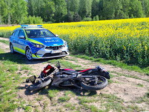 Zdjęcie przedstawia oznakowany radiowóz policji, przed którym leży przewrócony motorower. Pojazdy znajdują się na drodze gruntowej.