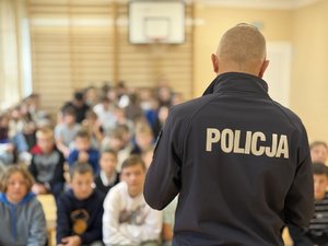 Zdjęcia przedstawiają umundurowanego funkcjonariusza policji podczas pogadanki w szkołach. W tle widać dzieci. Policjant stoi tyłem do obiektywu aparatu.