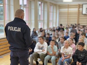 Zdjęcia przedstawiają umundurowanego funkcjonariusza policji podczas pogadanki w szkołach. W tle widać dzieci. Policjant stoi tyłem do obiektywu aparatu.