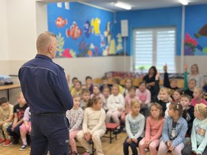 Zdjęcia przedstawiają umundurowanego funkcjonariusza policji podczas pogadanki w szkołach. W tle widać dzieci. Policjant stoi tyłem do obiektywu aparatu.