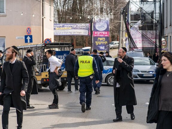 Zdjęcie przedstawia umundurowanego funkcjonariusza policji, który odwrócony jest tyłem do obiektywu. Dookoła niego widać chodzących pielgrzymów.
